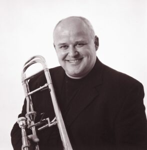 Svart/hvitt bilde av PK Svensen, trombonist og professor, lener trombonen sin på høyre skulder.